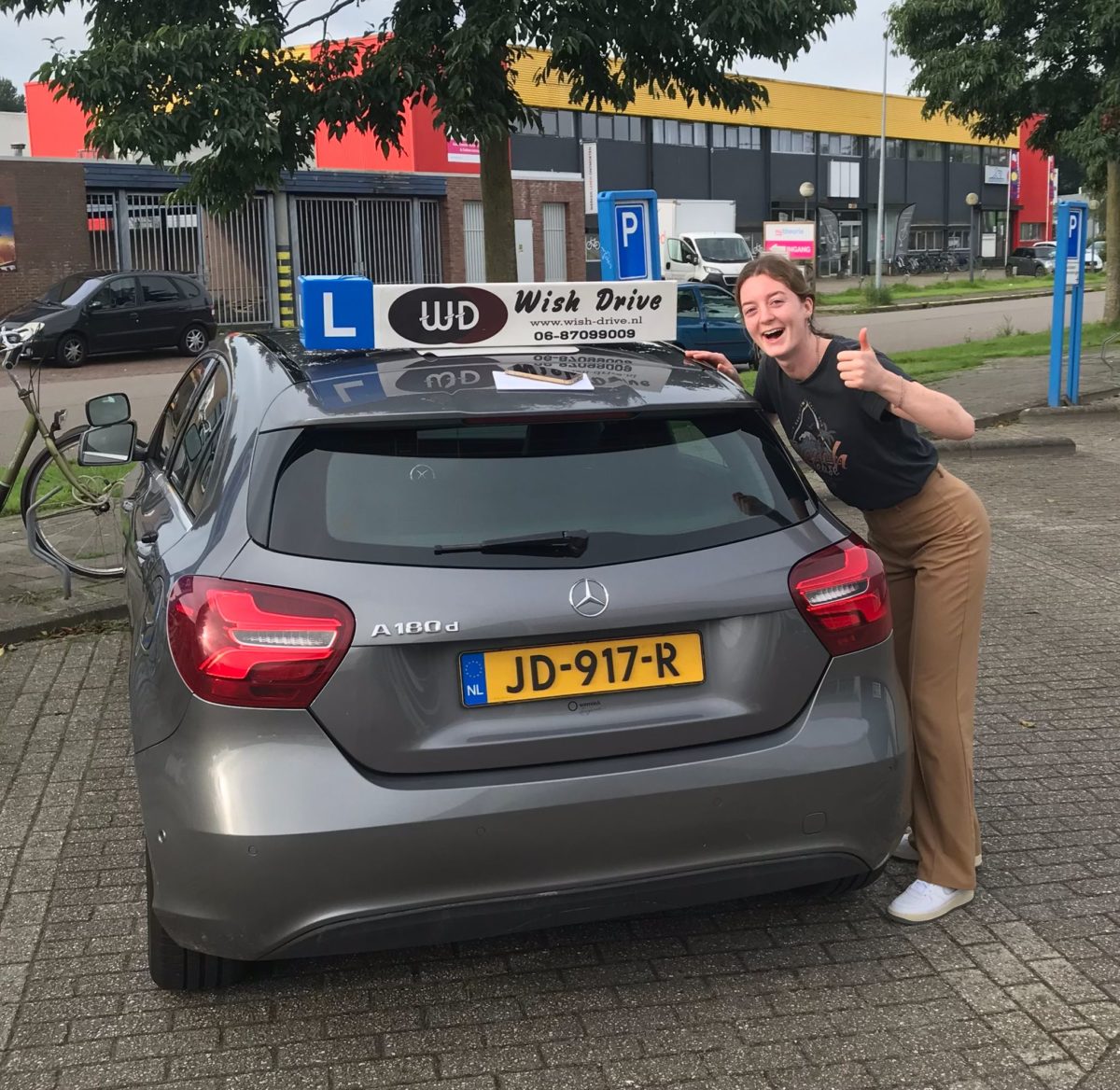 Geslaagde leerling poseert blij naast de lesauto van Wish Drive